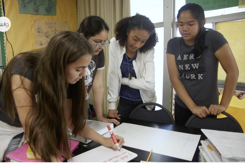 Four students around a desk writing on a poster.