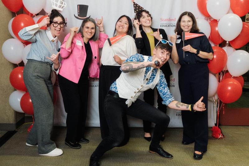 Six event participants pose festively for a photo against a backdrop with balloons